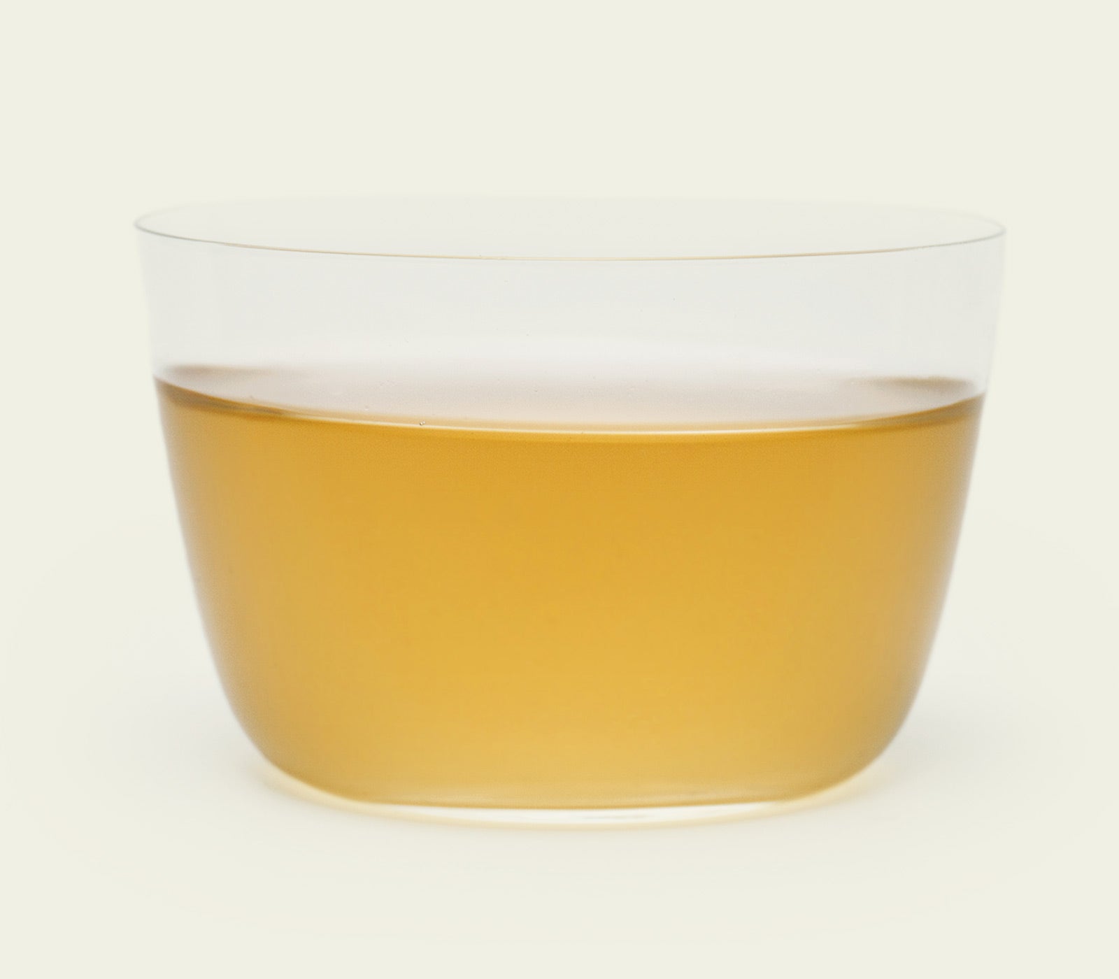 Mao Feng Tee – Seitliche Aufnahme eines klaren Glases, das den hellgoldenen Farbton des Tees zeigt, mit einem sanften Übergang in die Tassenfarbe.